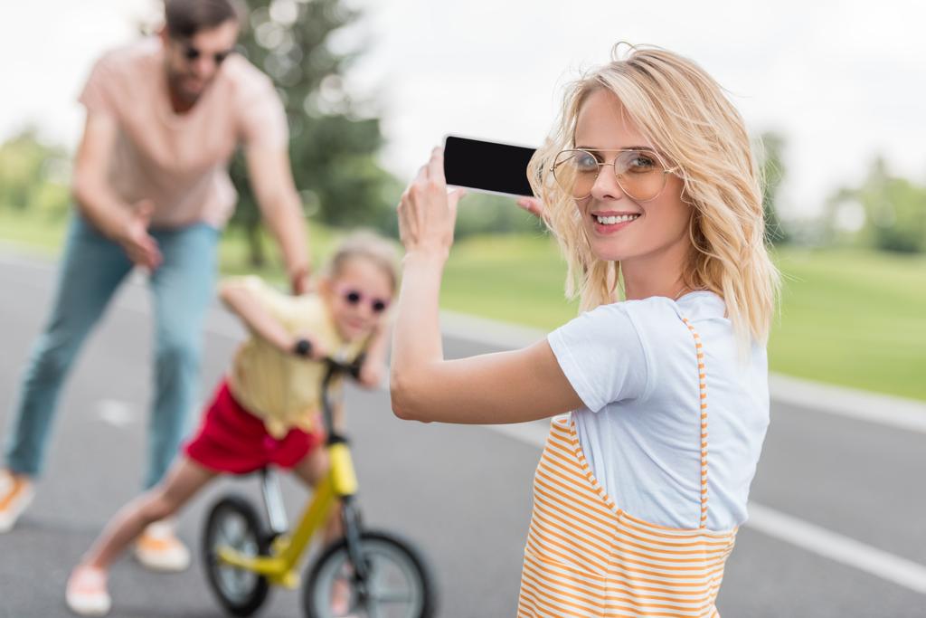 woman holding smartphone and smiling at camera while photographing father teaching daughter riding bicycle - Photo, Image