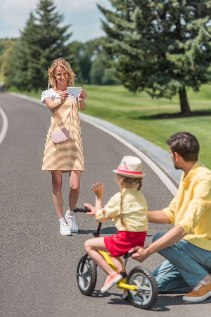 щаслива молода жінка зі смартфоном фотографує батька навчаючи дочку їздити на велосипеді в парку
 - Фото, зображення