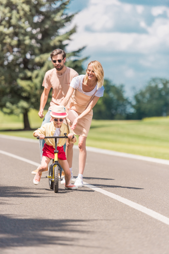 šťastné mladé rodiče při pohledu na malou dceru jedoucí na kole v parku  - Fotografie, Obrázek