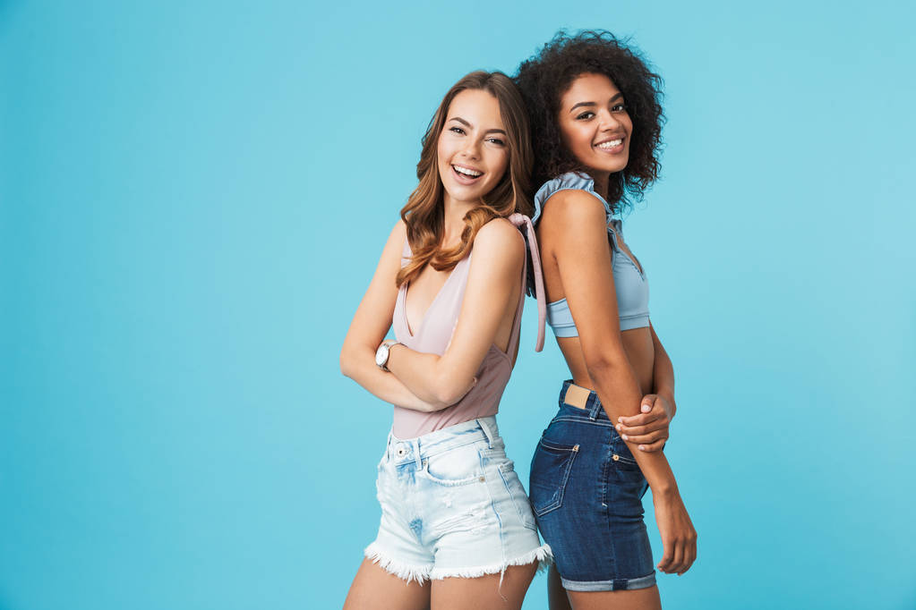 Dwie szczęśliwe młode dziewczyny ubrane w ubrania letnie, powrotem do tyłu stojąc i patrząc na kamery na białym tle niebieski - Zdjęcie, obraz