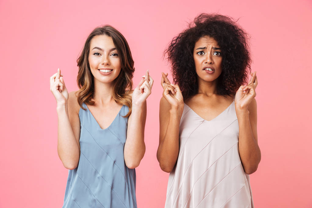 Dos jóvenes preocupadas vestidas con ropa de verano sosteniendo los dedos cruzados para la buena suerte aisladas sobre fondo rosa
 - Foto, Imagen