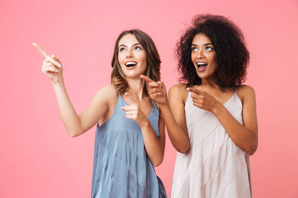 Две счастливые девушки, одетые в летнюю одежду, указывают на пространство для копирования, изолированное на розовом фоне.
 - Фото, изображение