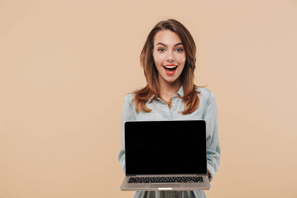 Portret van een enthousiast jong meisje in zomer kleding toont leeg scherm laptopcomputer geïsoleerd op beige achtergrond - Foto, afbeelding
