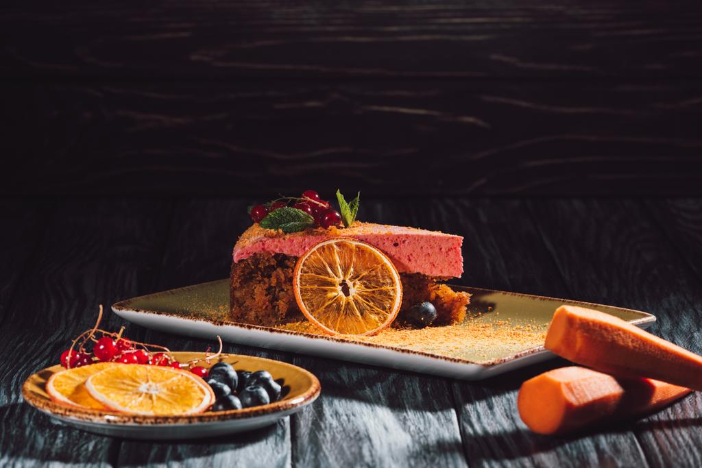 κλείνω πάνω θέα του κέικ καρότου με τη μέντα και πλήρωση μούρων, καρότα, φέτες πορτοκαλιού και τα μούρα σχετικά με πιατάκι στο ξύλινο τραπέζι  - Φωτογραφία, εικόνα