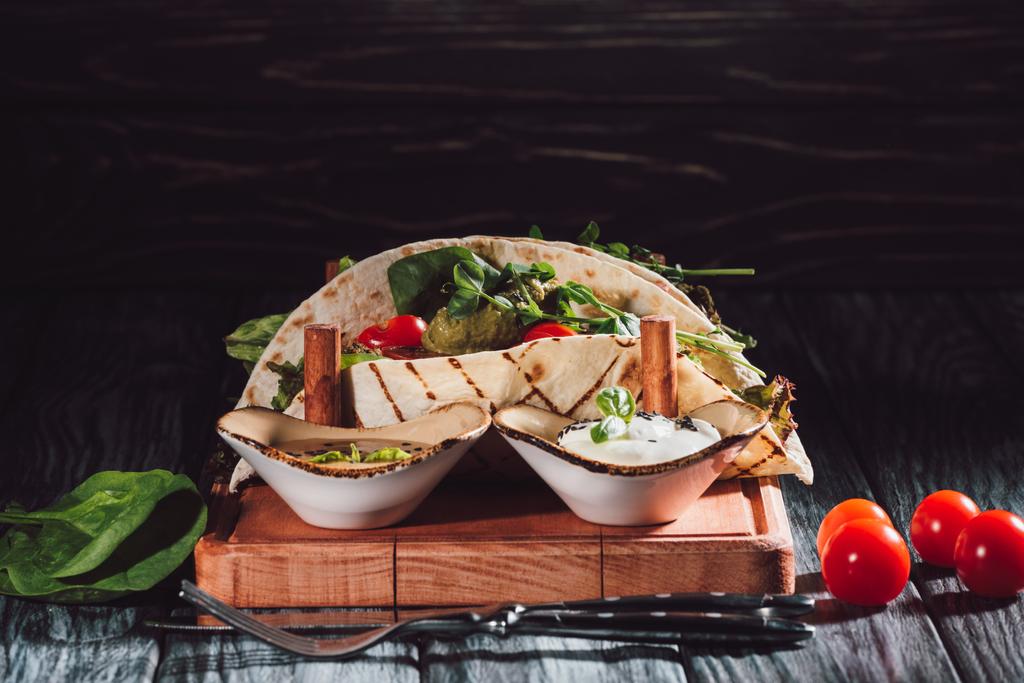 Close-up van tortilla's met falafel, cherrytomaatjes en gekiemde zaden van zonnebloem op houten dienblad met sauzen in de buurt van de vork en mes op houten tafel  - Foto, afbeelding