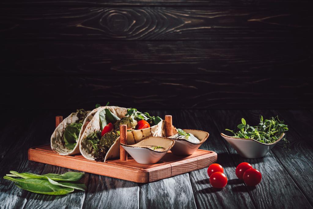 composición alimenticia de tortillas con falafel, tomates cherry y semillas germinadas de girasol en bandeja de madera con salsas sobre mesa
 - Foto, imagen