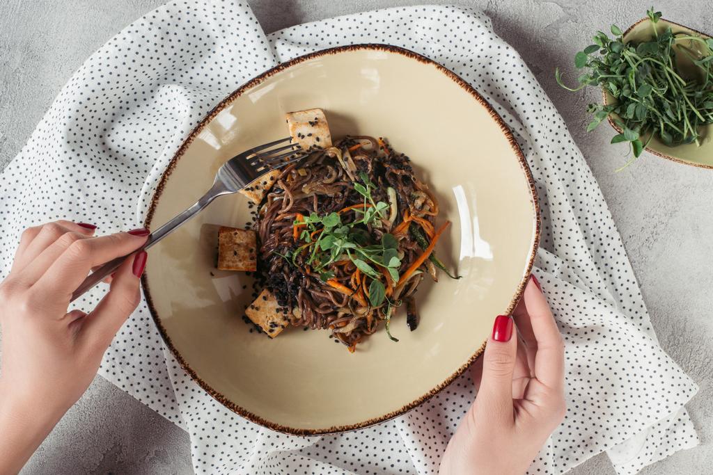partial view of female hands and soba with tofu and vegetables decorated with germinated seeds of sunflower on grey tabletop - Photo, Image