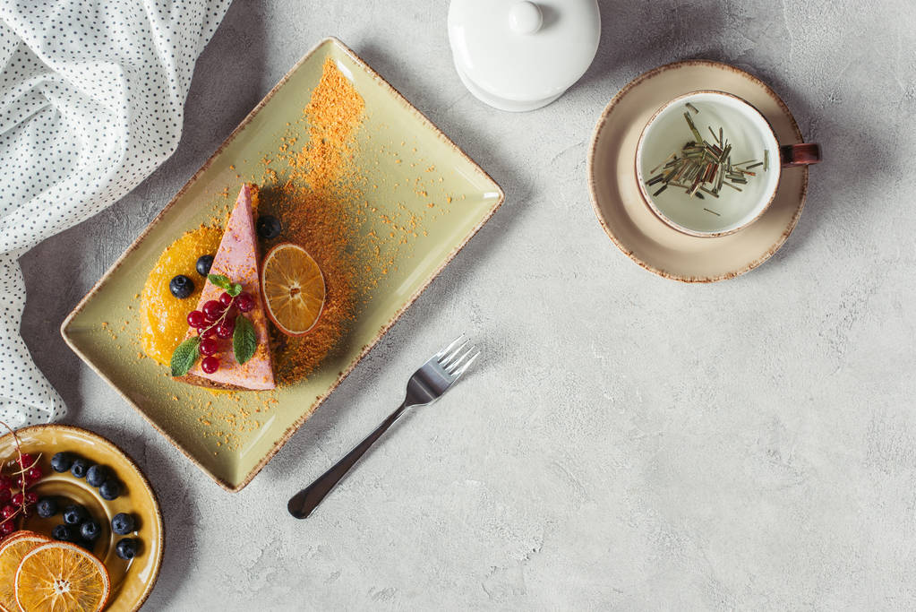 acostado plano con trozo de pastel de zanahoria dulce con relleno de bayas, taza de té de hierbas, ropa de cama y tenedor en la mesa gris
 - Foto, Imagen