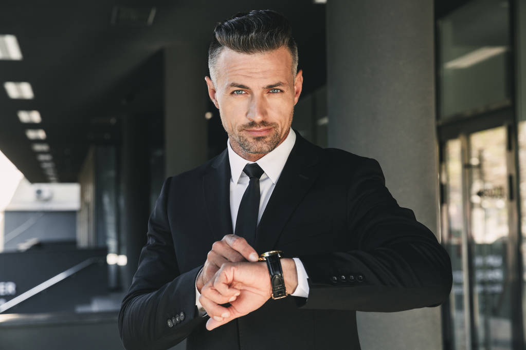 Portrait d'un jeune homme d'affaires confiant habillé en costume debout à l'extérieur d'un bâtiment en verre et regardant sa montre-bracelet
 - Photo, image