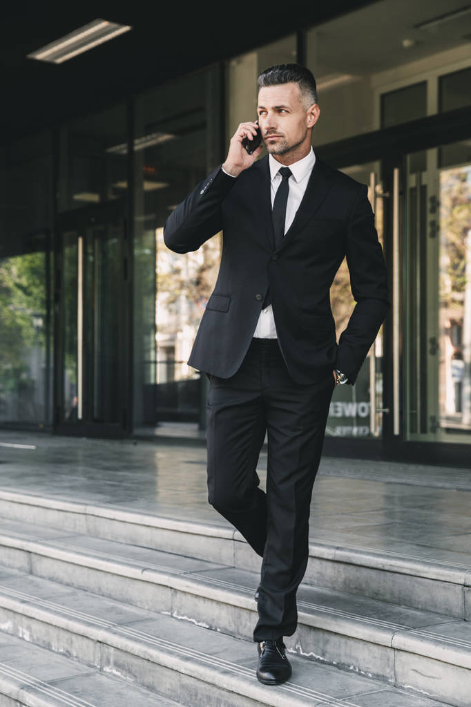 Muotokuva komea nuori liikemies puvussa kävelee alas portaita lasirakennuksen ulkopuolella ja puhuu matkapuhelimella
 - Valokuva, kuva