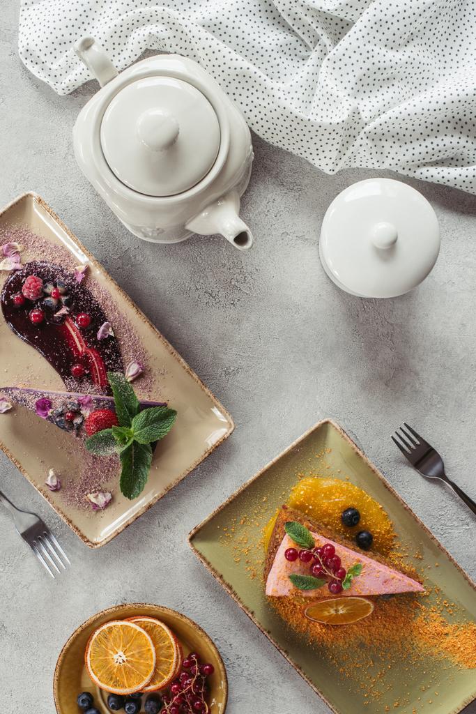 ベリーいっぱいの甘いにんじんケーキとフラット レイアウトは、ブルーベリーのケーキを添えてミントの葉と紫の花びら、ティーポット、灰色の卓上のリネン - 写真・画像