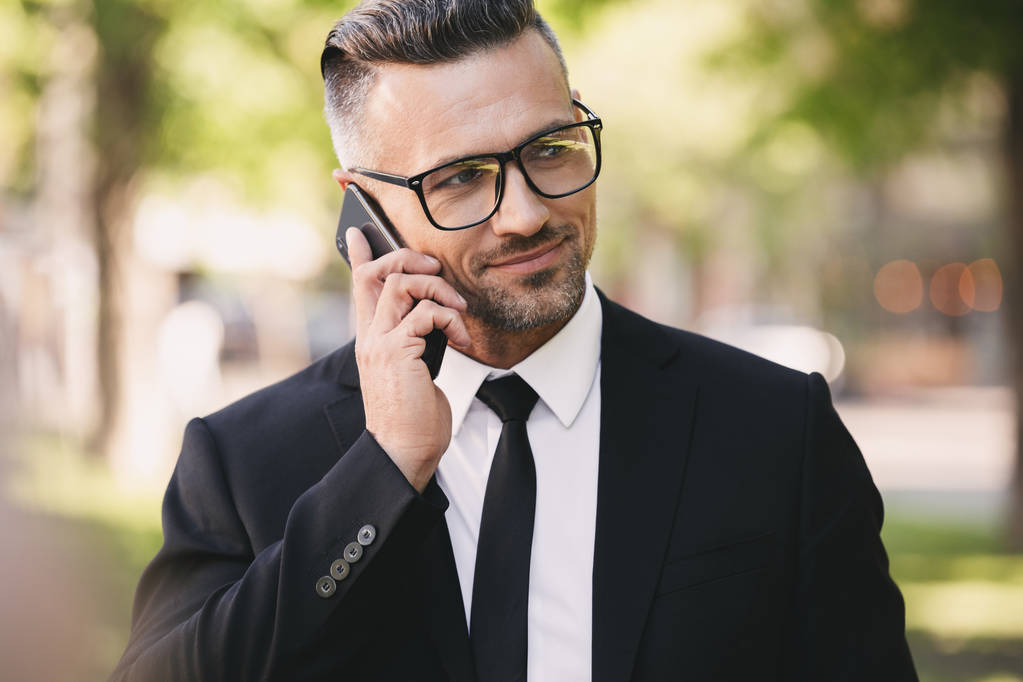 Портрет счастливого бизнесмена в костюме, идущего на улицу и пользующегося мобильным телефоном
 - Фото, изображение