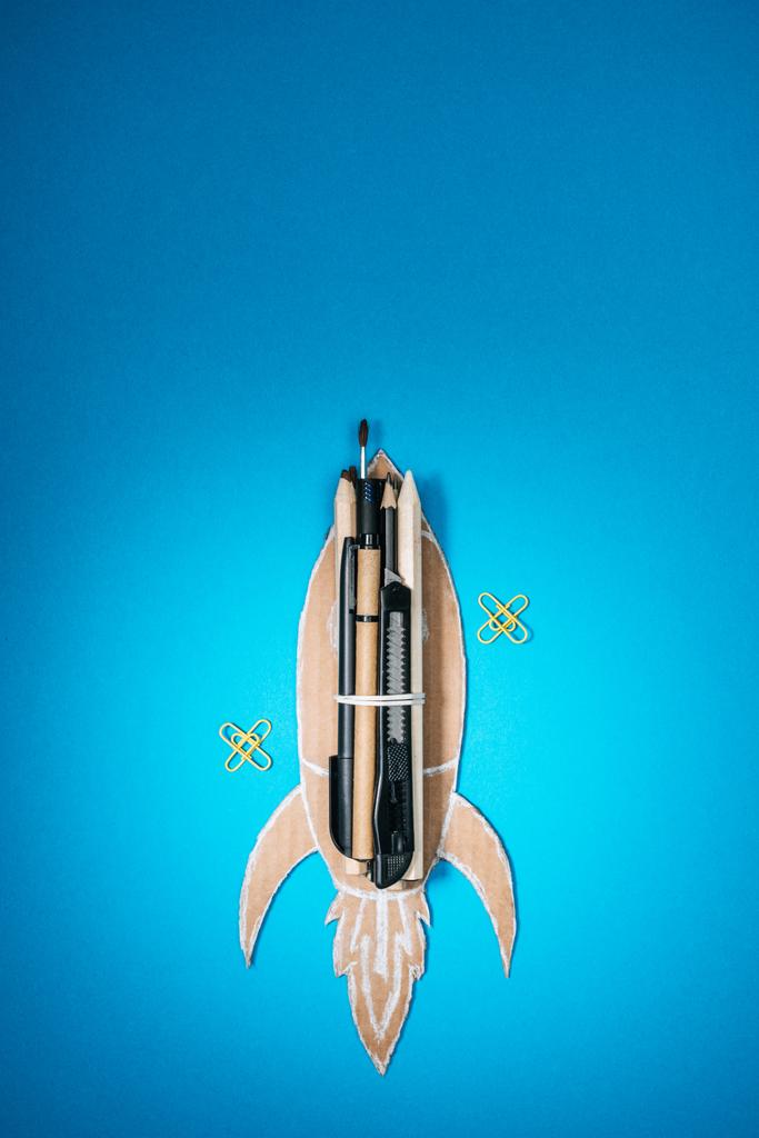 вид сверху на картонную ракету ручной работы с канцелярскими принадлежностями и скрепками на синем фоне
 - Фото, изображение
