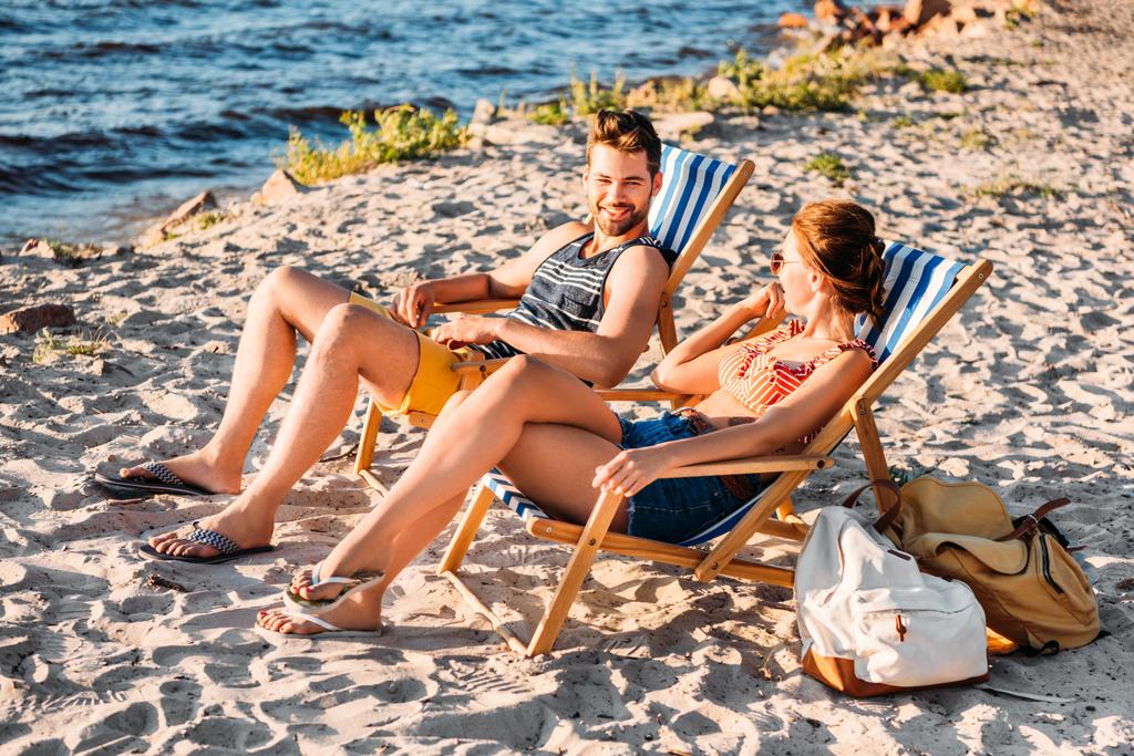 vista de alto ângulo de feliz jovem casal descansando em salões de chaise na praia de areia
 - Foto, Imagem