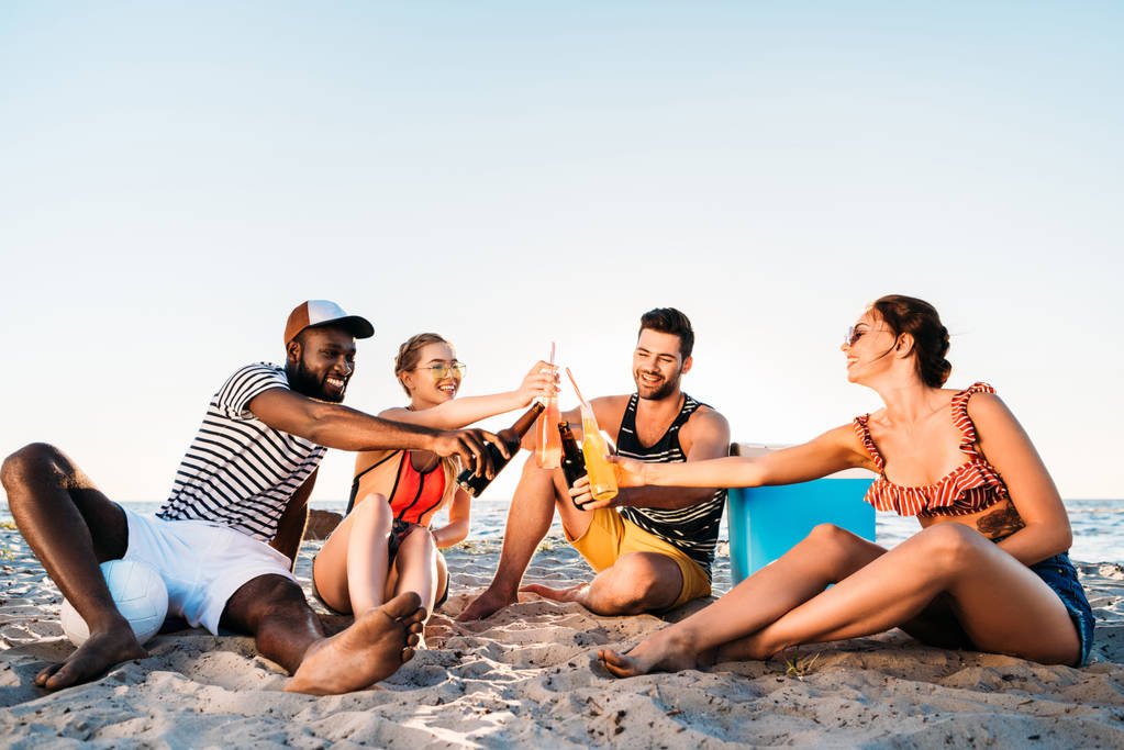 szczęśliwy młodzi przyjaciele wieloetnicznym brzęk szkła butelki z napojów siedząc razem na plaży  - Zdjęcie, obraz