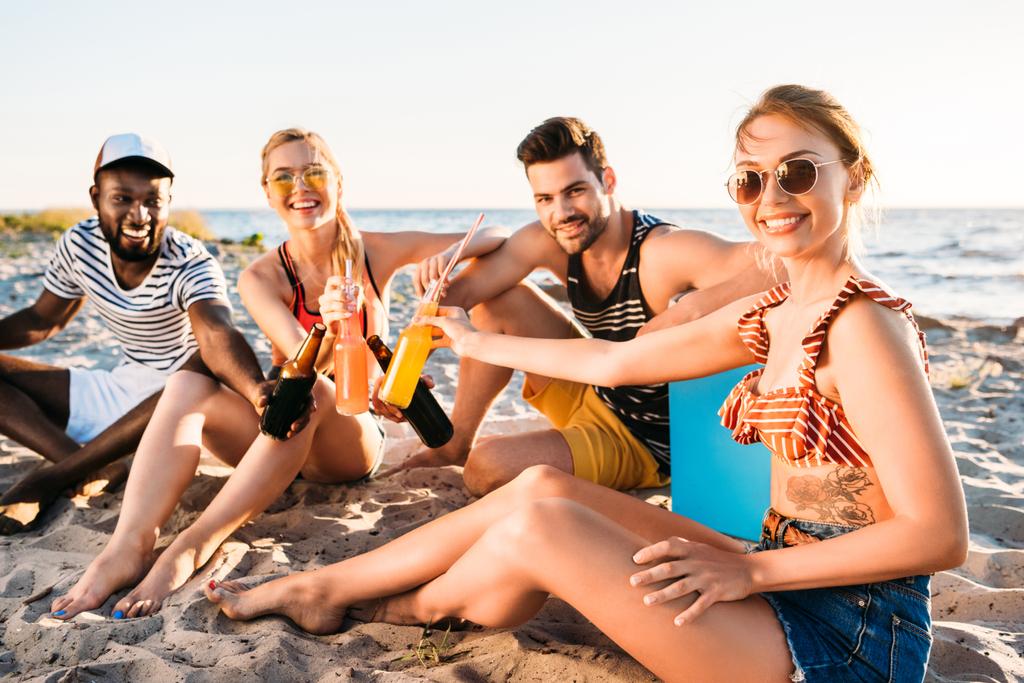 heureux jeunes amis multiethniques tenant des bouteilles en verre avec des boissons et souriant à la caméra sur la plage de sable
 - Photo, image