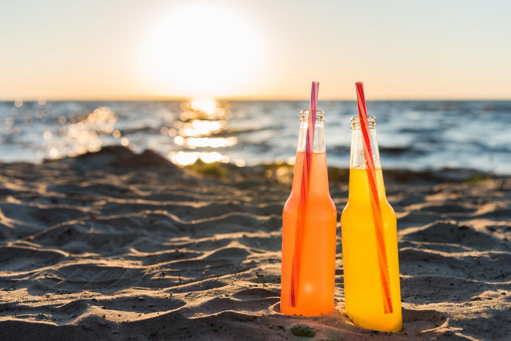 Nahaufnahme von Glasflaschen mit Erfrischungsgetränken und Trinkhalmen am Sandstrand bei Sonnenuntergang - Foto, Bild
