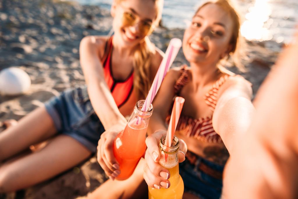 επιλεκτική εστίαση του ευχαριστημένοι νέοι φίλες κρατώντας γυάλινα μπουκάλια με ποτά το καλοκαίρι στην παραλία - Φωτογραφία, εικόνα