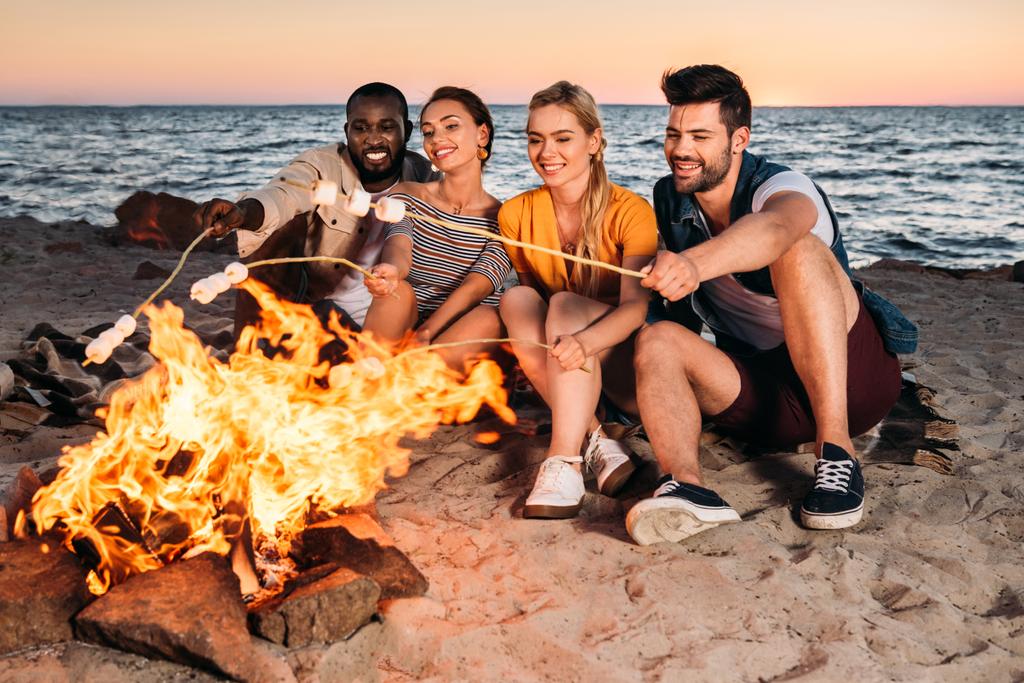 happy young multiethnic friends roasting marshmallows at bonfire on sandy beach at sunset  - Photo, Image