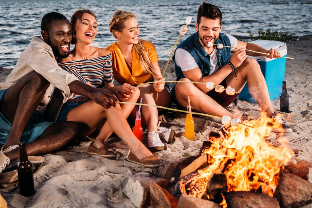 Happy mladých mnohonárodnostní přátelé, pražení marshmallows na táborák na pláži při západu slunce  - Fotografie, Obrázek