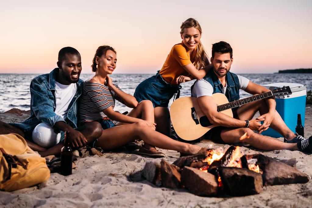 happy young multiethnic friends with guitar looking at bonfire while sitting together on sandy beach - Photo, Image