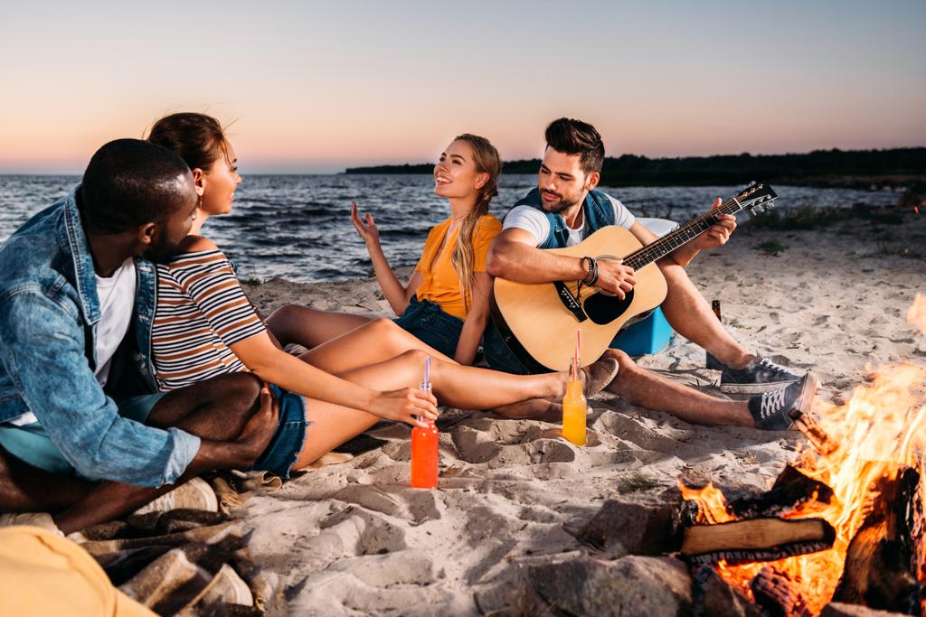 boldog fiatal többnemzetiségű meg élvezik a gitár, és tölteni az időt együtt a homokos tengerparton naplementekor  - Fotó, kép