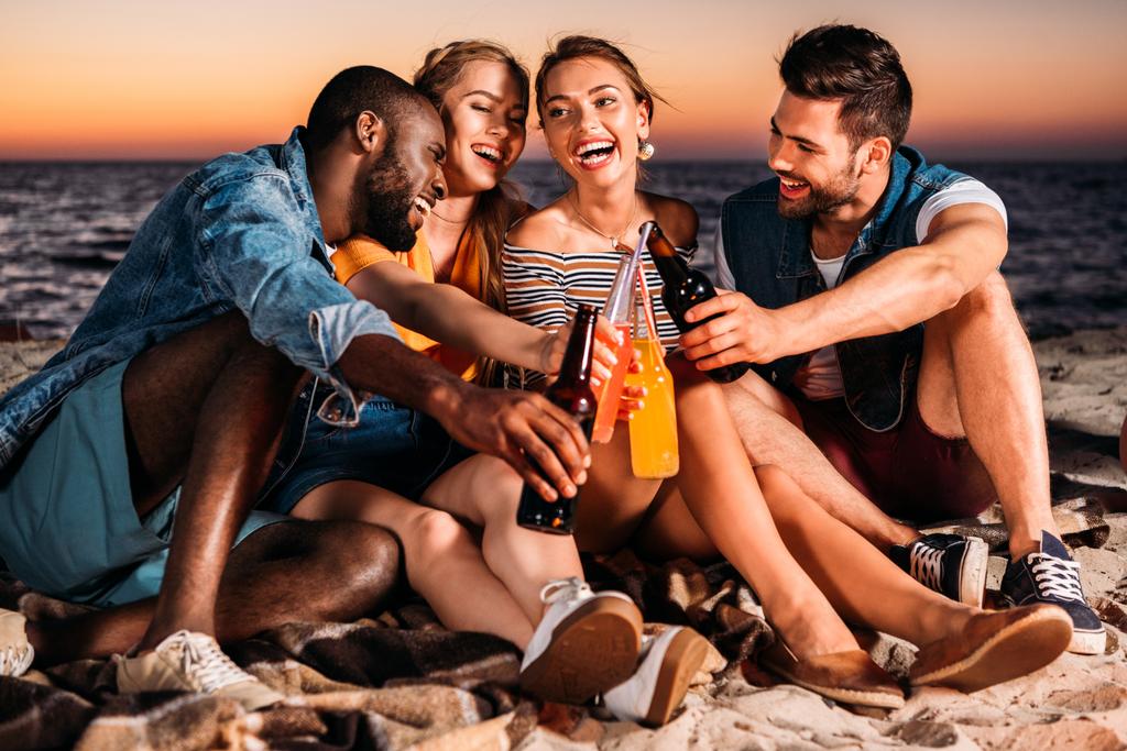 jóvenes felices tintineando botellas de vidrio con bebidas mientras pasan tiempo juntos en la playa de arena al atardecer
  - Foto, imagen