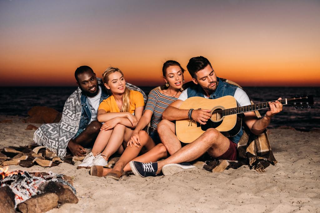 happy young multiethnic friends enjoying guitar on beach at sunset - Photo, Image