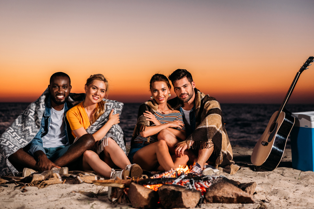 happy young people smiling at camera while sitting near bonfire on sandy beach at sunset  - Photo, Image