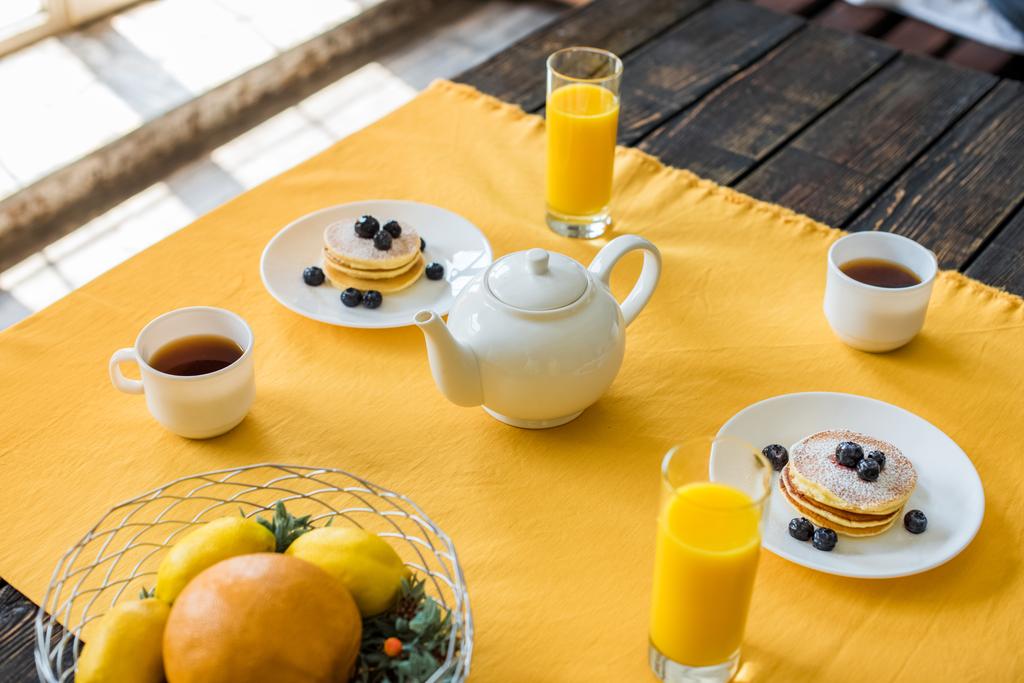 vista da vicino di frittelle, tazze di tè e bicchieri di succo di frutta per la colazione sul tavolo con tovaglia gialla
 - Foto, immagini