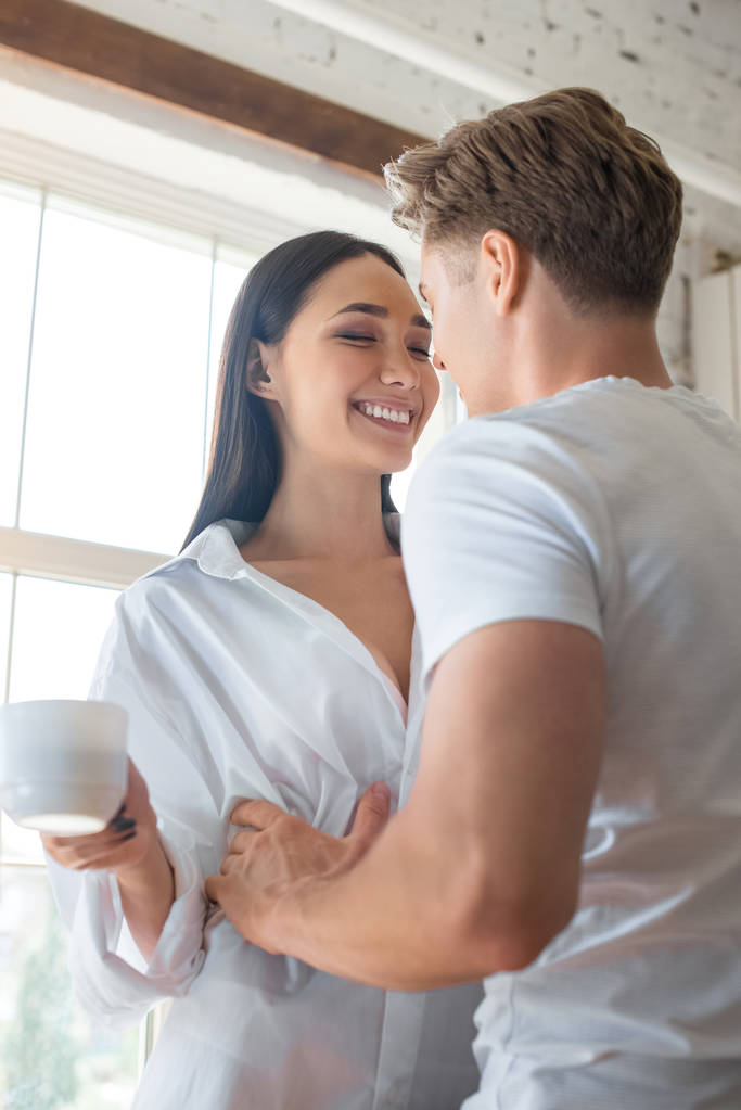 caucásico novio abrazando sonriente asiático mujer con taza de café en casa
 - Foto, imagen