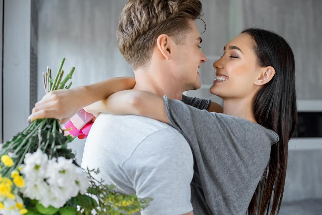 ギフトやブーケの花と家でお互いをハグ白人のボーイ フレンドを持つ幸せなアジア女の側面図 - 写真・画像