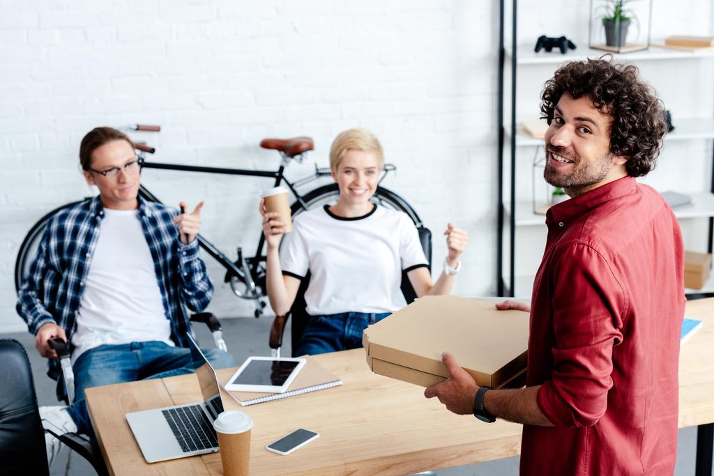 uśmiechający się młodych kolegów ogląda szczęśliwy człowiek posiadający pudełek po pizzy w biurze  - Zdjęcie, obraz