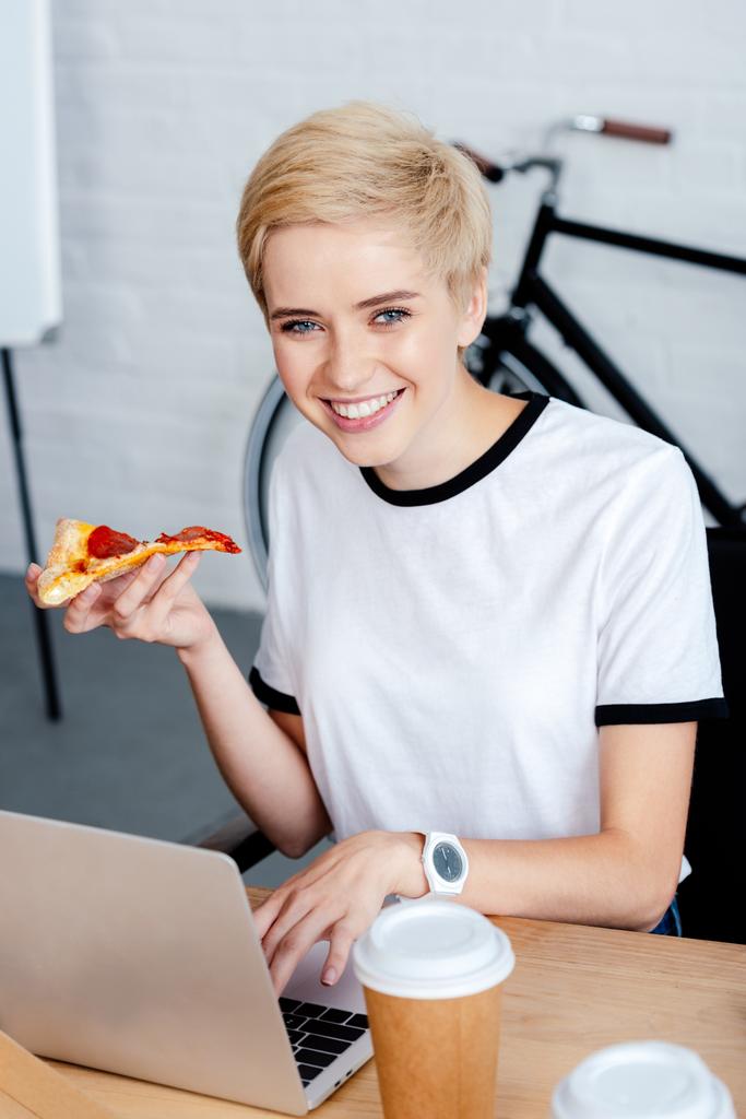 hoge hoekmening van meisje met pizza met behulp van laptop en lachend op camera - Foto, afbeelding