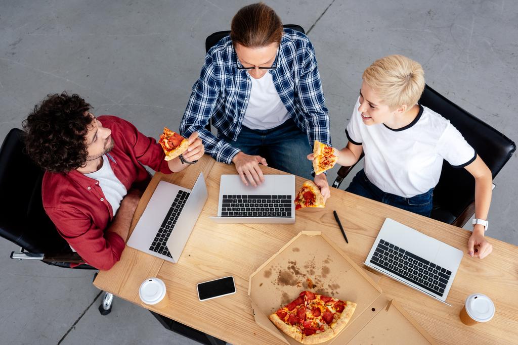 вид сверху на молодых коллег, которые едят пиццу и используют ноутбуки на рабочем месте
 - Фото, изображение