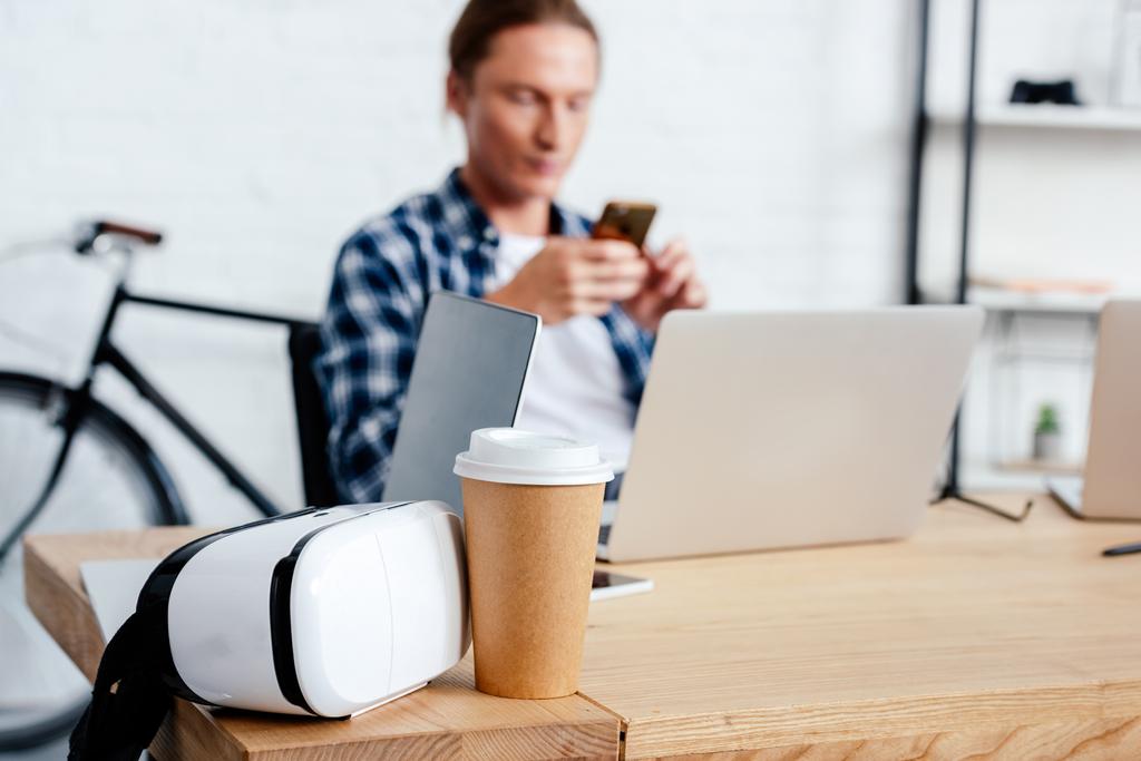 vista de cerca de café para llevar, auriculares de realidad virtual y el hombre utilizando el teléfono inteligente detrás
 - Foto, imagen