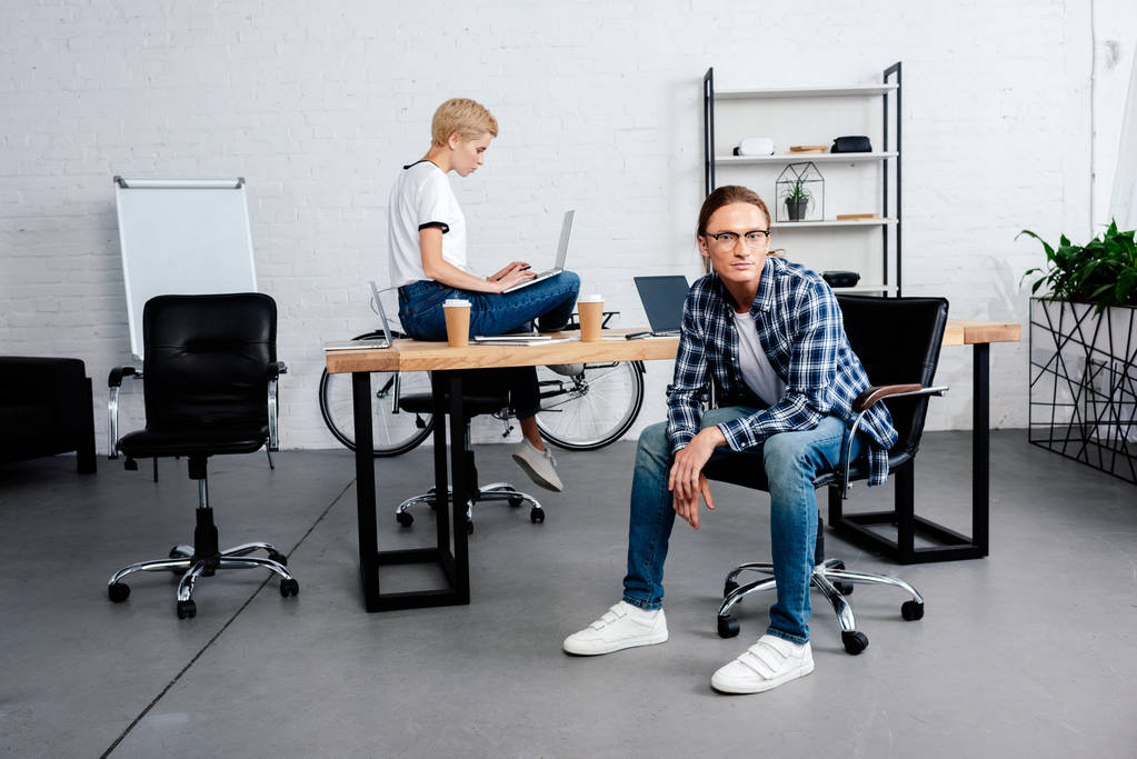 mujer joven usando el ordenador portátil y colega masculino mirando a la cámara en la oficina
 - Foto, imagen