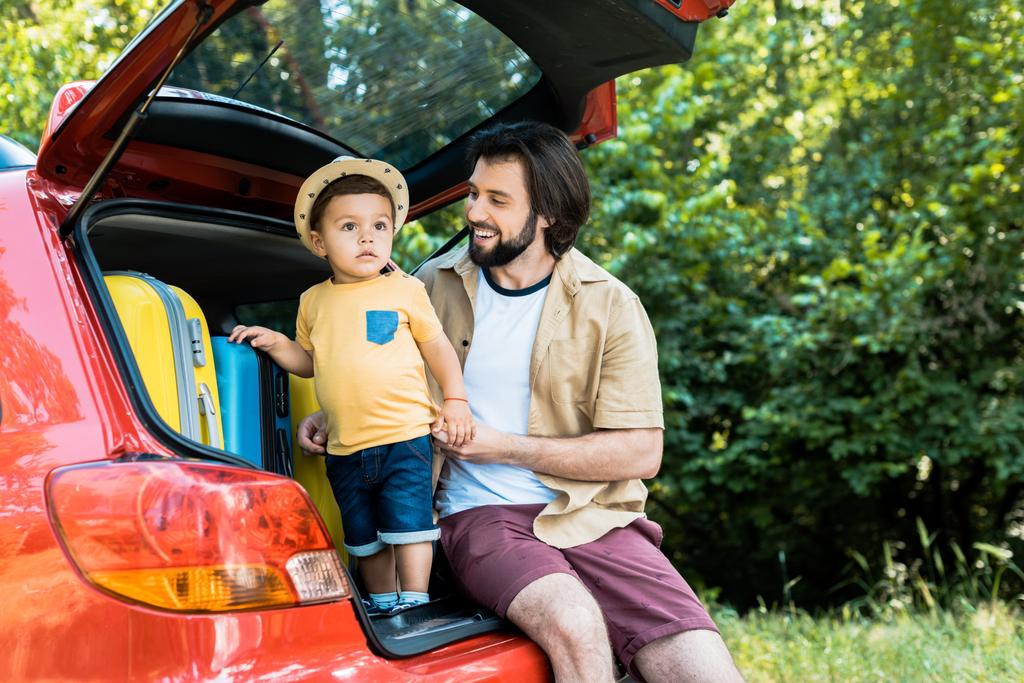 padre e figlio sorridenti seduti sul bagagliaio dell'auto nella foresta
 - Foto, immagini