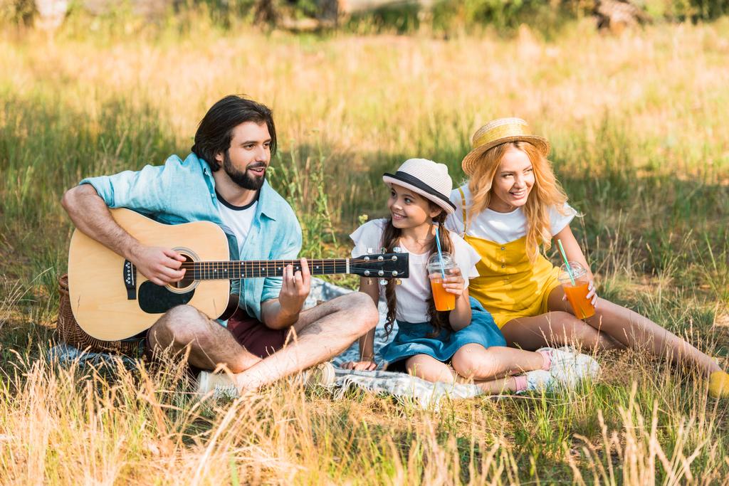 красивый отец играет на акустической гитаре для дочери и жены на пикнике
 - Фото, изображение