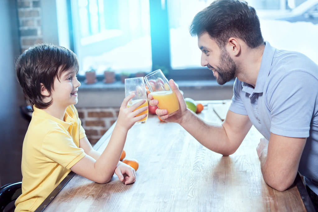 χαμογελώντας μικρό αγόρι και ο πατέρας του τσούγκριζαν από ποτήρια με χυμό στο τραπέζι  - Φωτογραφία, εικόνα