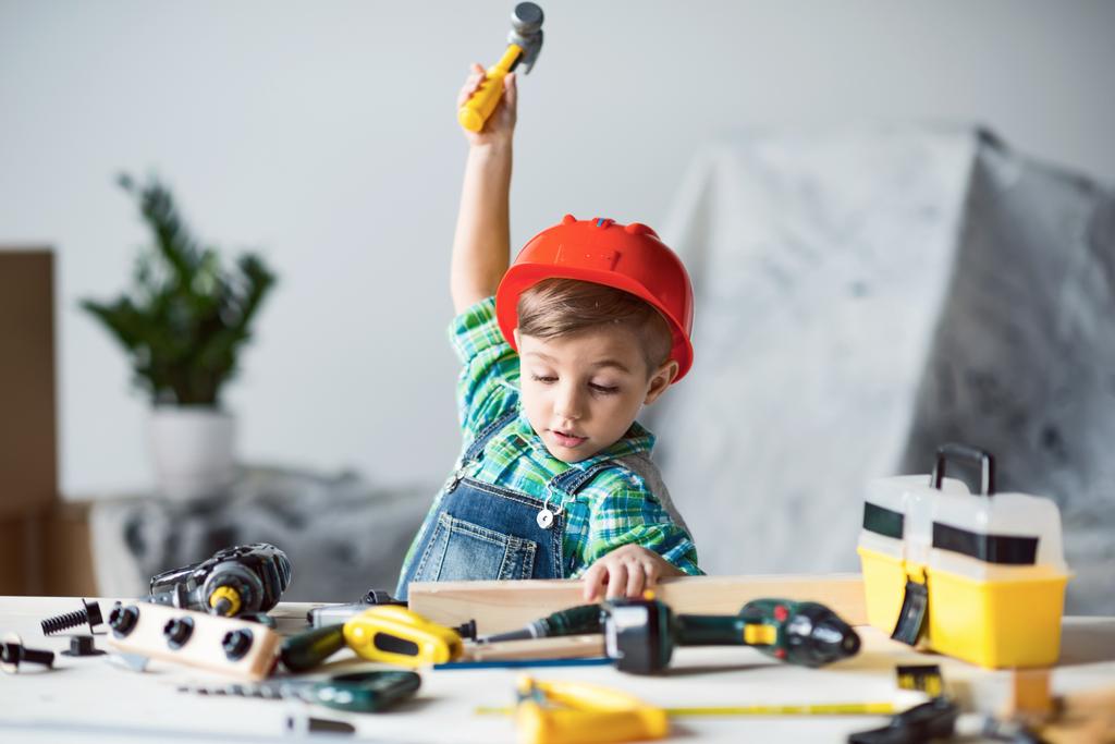 adorable little kid in red hard hat playing with wooden plank and toy hammer - Photo, Image
