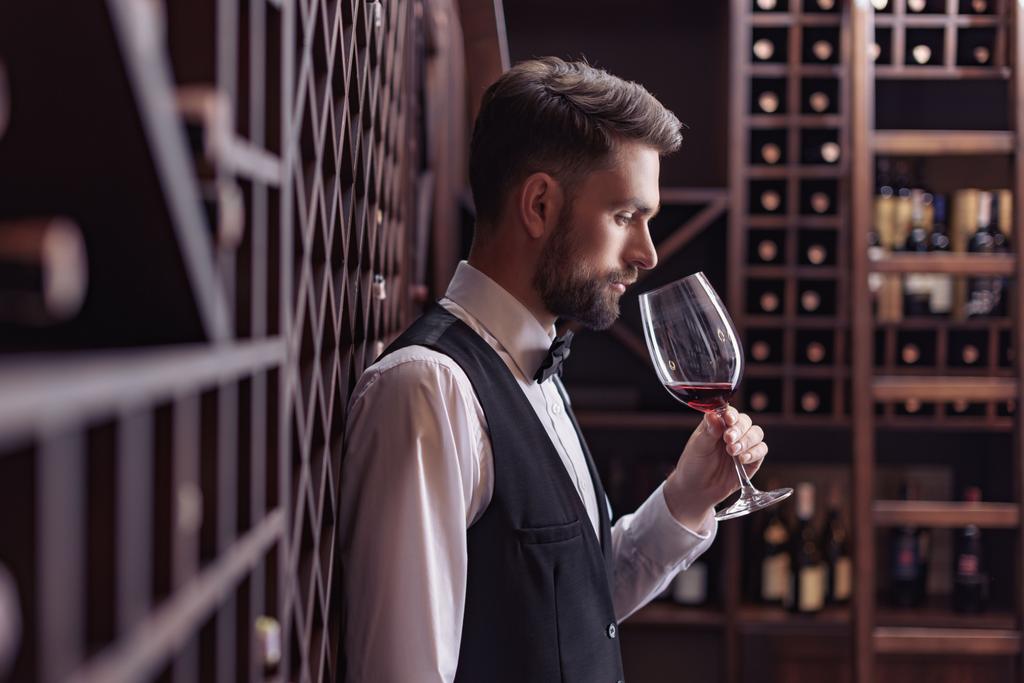 Seitenansicht eines jungen gutaussehenden Sommeliers, der Rotwein im Weinkeller verkostet, während er sich auf Regale lehnt - Foto, Bild