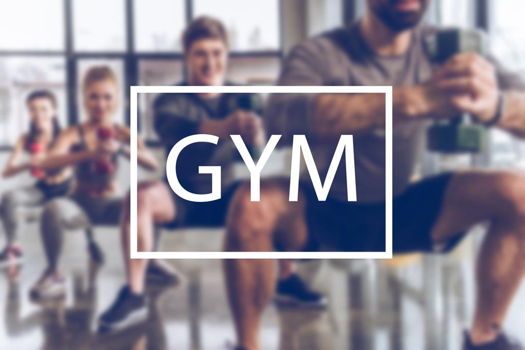 blurred group of athletic young people in sportswear with dumbbells exercising at gym, gym inscription - Photo, Image