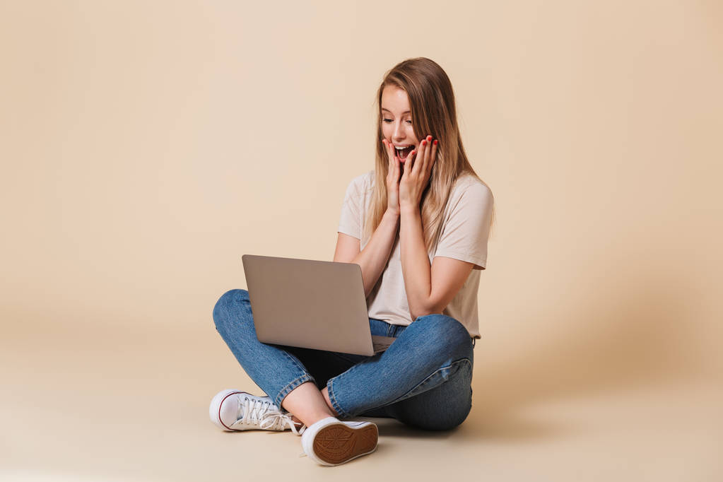 Portré egy izgatott alkalmi lány látszó-on laptop számítógép ülve, a lábak a padló elszigetelt átment bézs háttér - Fotó, kép