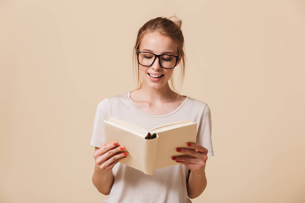 Πορτρέτο του ένα χαμογελαστό νεαρό κορίτσι σε γυαλιά διαβάζοντας ένα βιβλίο που απομονώνονται σε μπεζ φόντο - Φωτογραφία, εικόνα