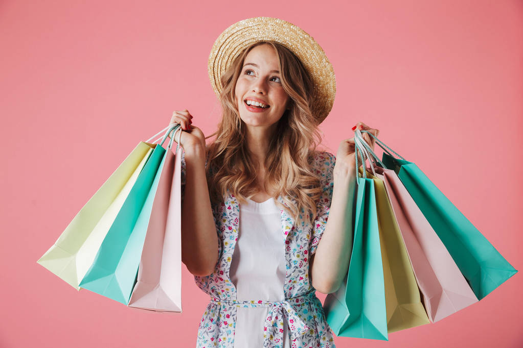 Portrait of a happy young woman in summer dress and straw hat holding shopping bags and looking away isolated over pink background - Photo, Image
