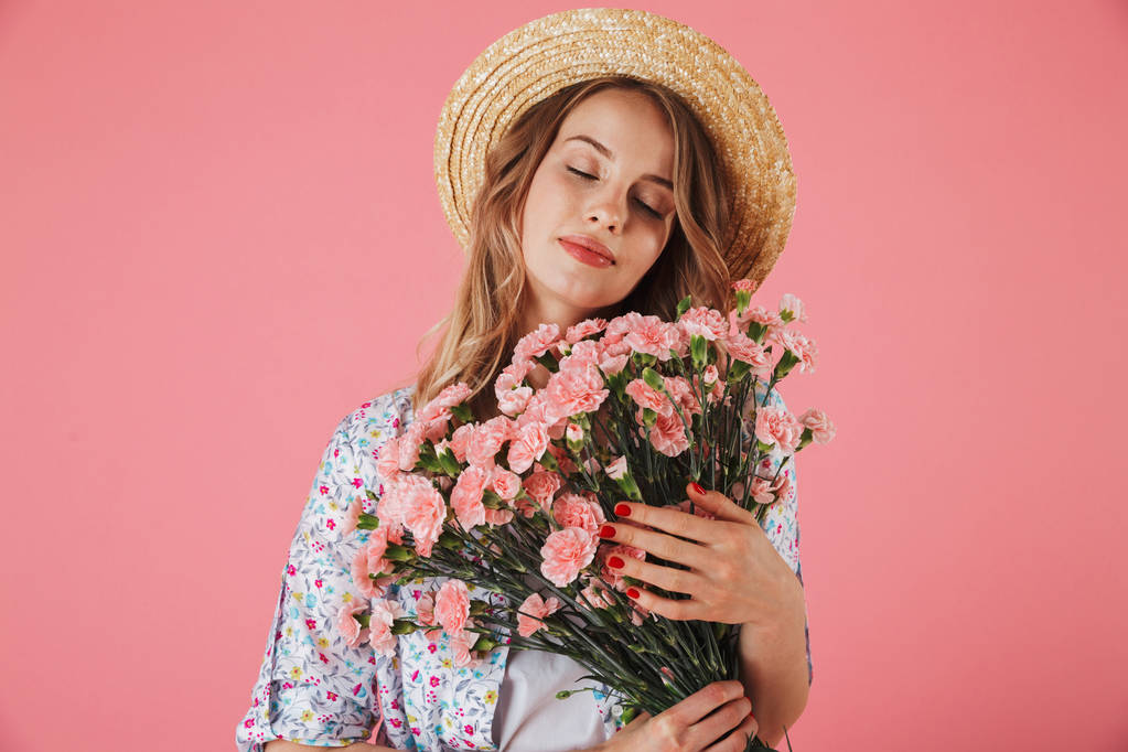 Portrait of a sensual young woman in summer dress and straw hat holding carnations bouquet isolated over pink background - Photo, Image