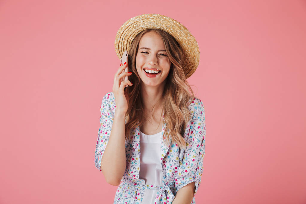 Retrato de una joven risueño en vestido de verano y sombrero de paja hablando en el teléfono móvil aislado sobre fondo rosa
 - Foto, imagen