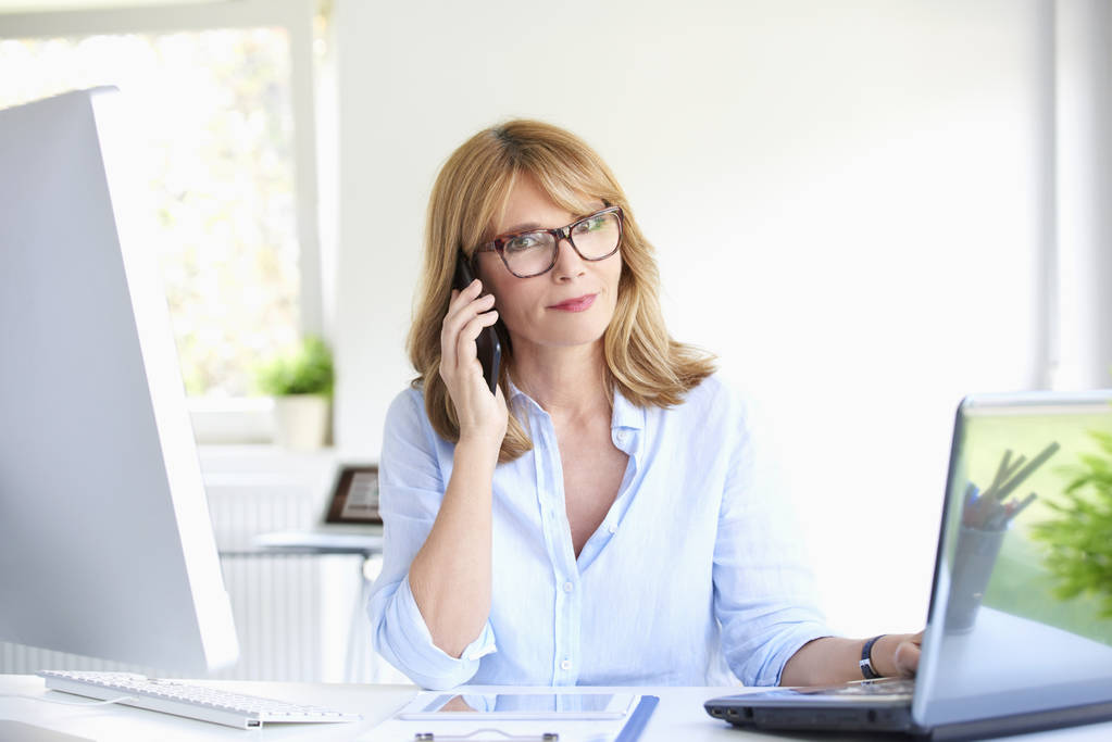 Крупним планом портрет привабливої зрілої бізнес-леді, яка розмовляє зі своїм клієнтом на мобільному телефоні, сидячи за офісним столом, працюючи на ноутбуці
. - Фото, зображення