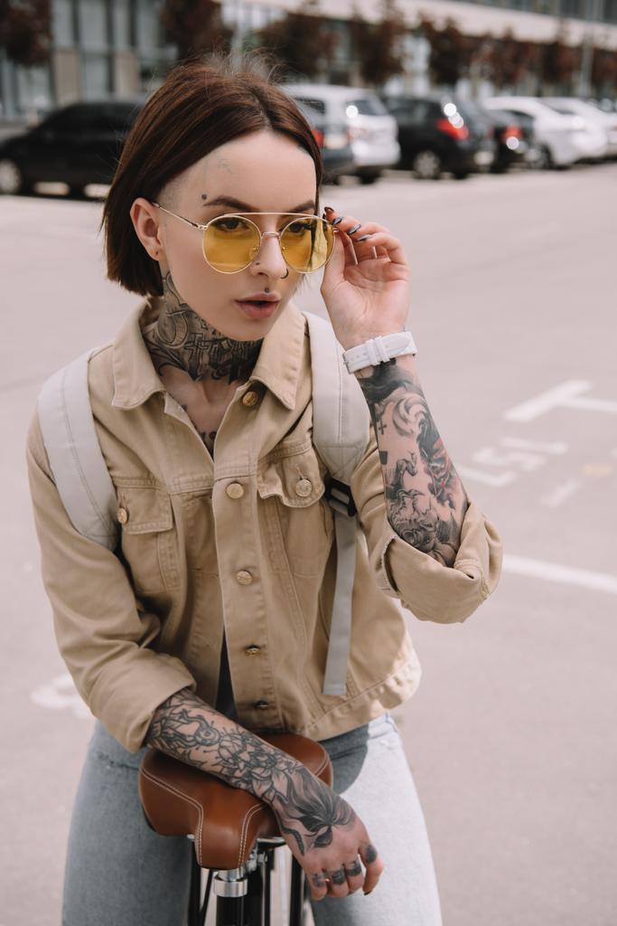 ελκυστική μοντέρνα τατουάζ γυναίκα να στέκεται με ποδήλατο στο δρόμο  - Φωτογραφία, εικόνα
